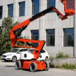 Оранжевый коленчатый подъемник с поднятой стрелой на фоне офисного здания и припаркованных машин.
